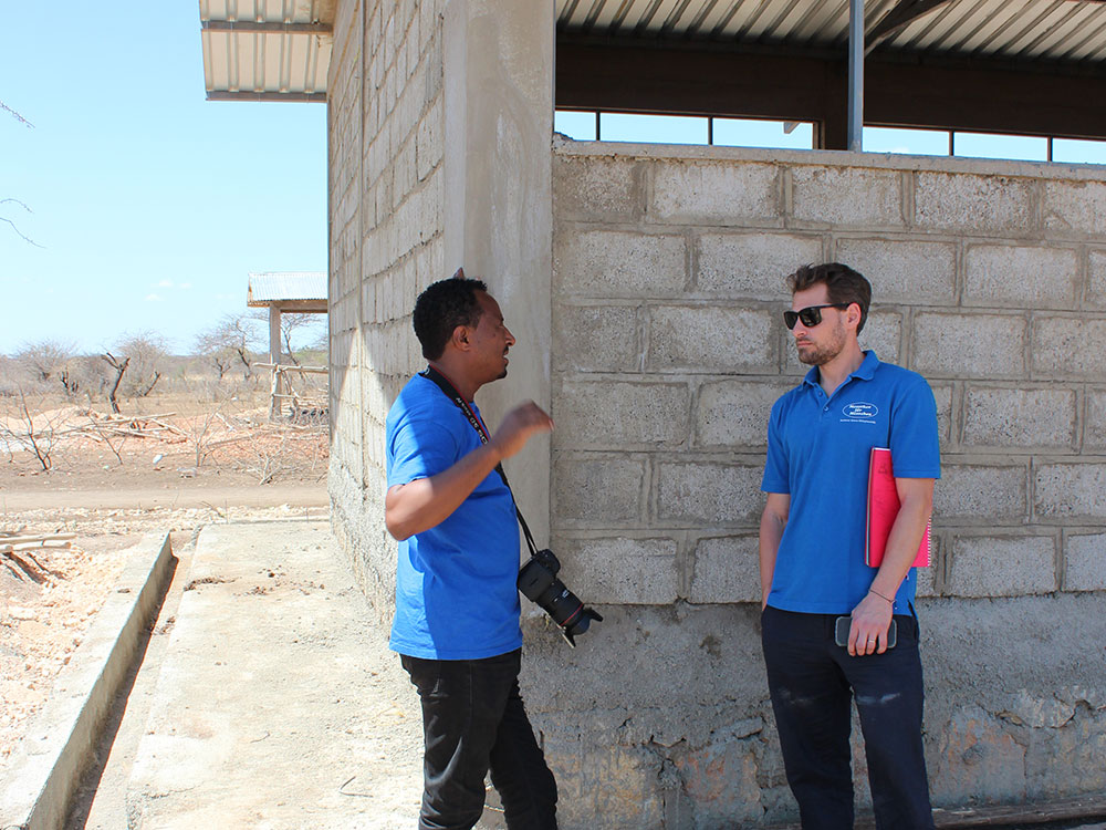 Muluneh Tolessa und Markus Schwarz-Herda tragen beide blaue Menschen für Menschen-Shirts und unterhalten sich an die Mauer eines Rohbaus einer Schule gelehnt.