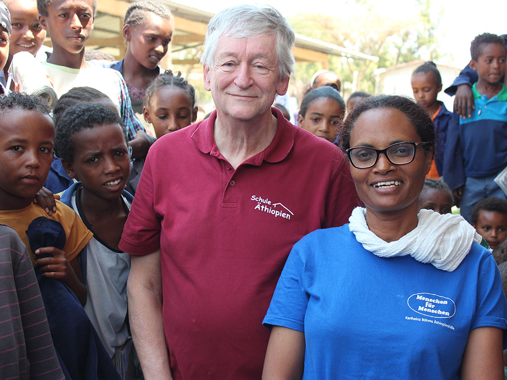 Peter Krasser in einem roten „Schule Äthiopien“-Shirt und Bahritu Seyoum in einem blauen „Menschen für Menschen“-Shirt stehen inmitten von Schulkindern. 