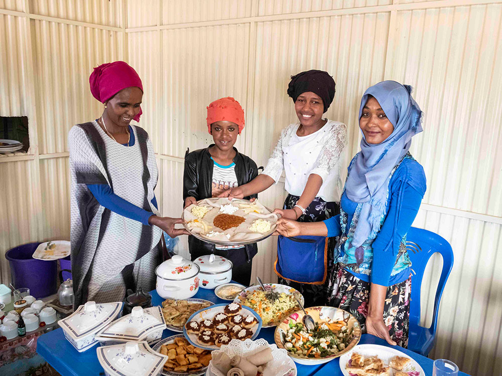 Vier junge äthiopische Frauen stehen um einen Tisch voll Speisen und präsentieren ein Teller auf dem ein Sauerteigfladen und Frischkäse ist