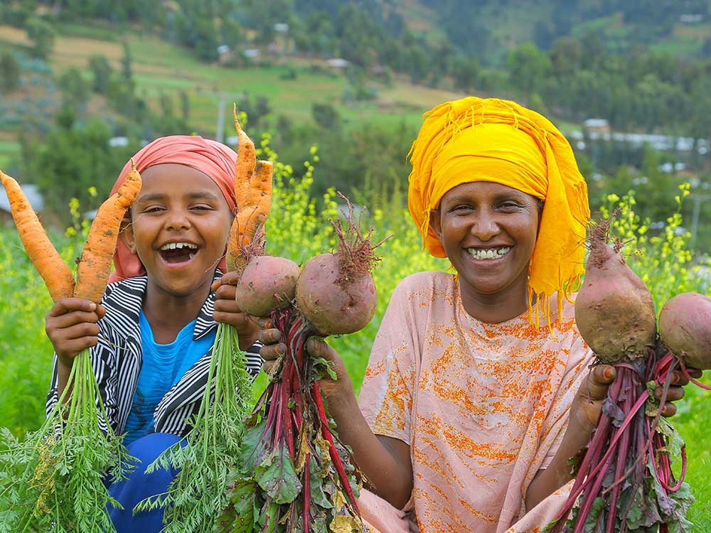 Eine Mutter und ihre kleine Tochter sitzen in einem Feld und lachen in die Kamera. Die Mutter hält rote Rüben in den Händen, die Tochter Karotten.