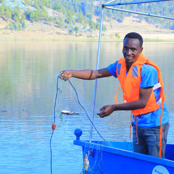 Ein Mann mit einer orangefarbenen Schwimmweste steht in einem blauen Boot und hält einen Fisch in einem Netz.
