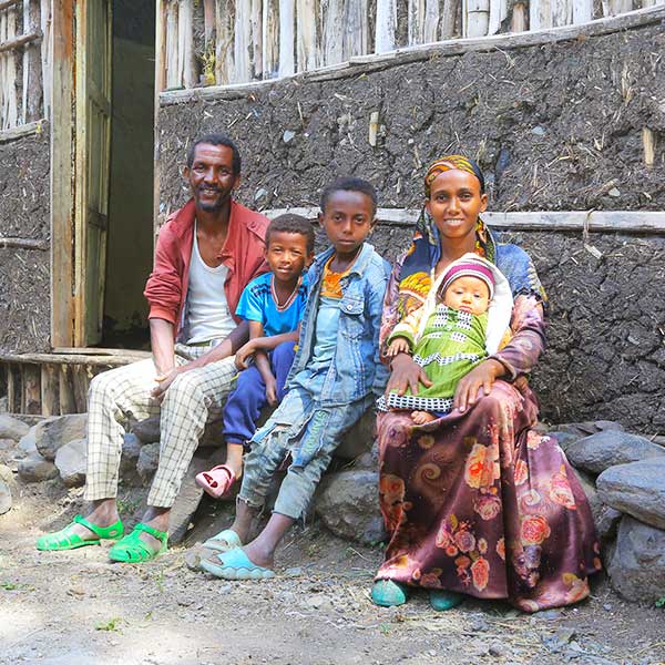 Eine Familie mit zwei Erwachsenen und drei Kindern sitzt vor einem einfachen Haus mit einer mit Lehm verputzten Wand.