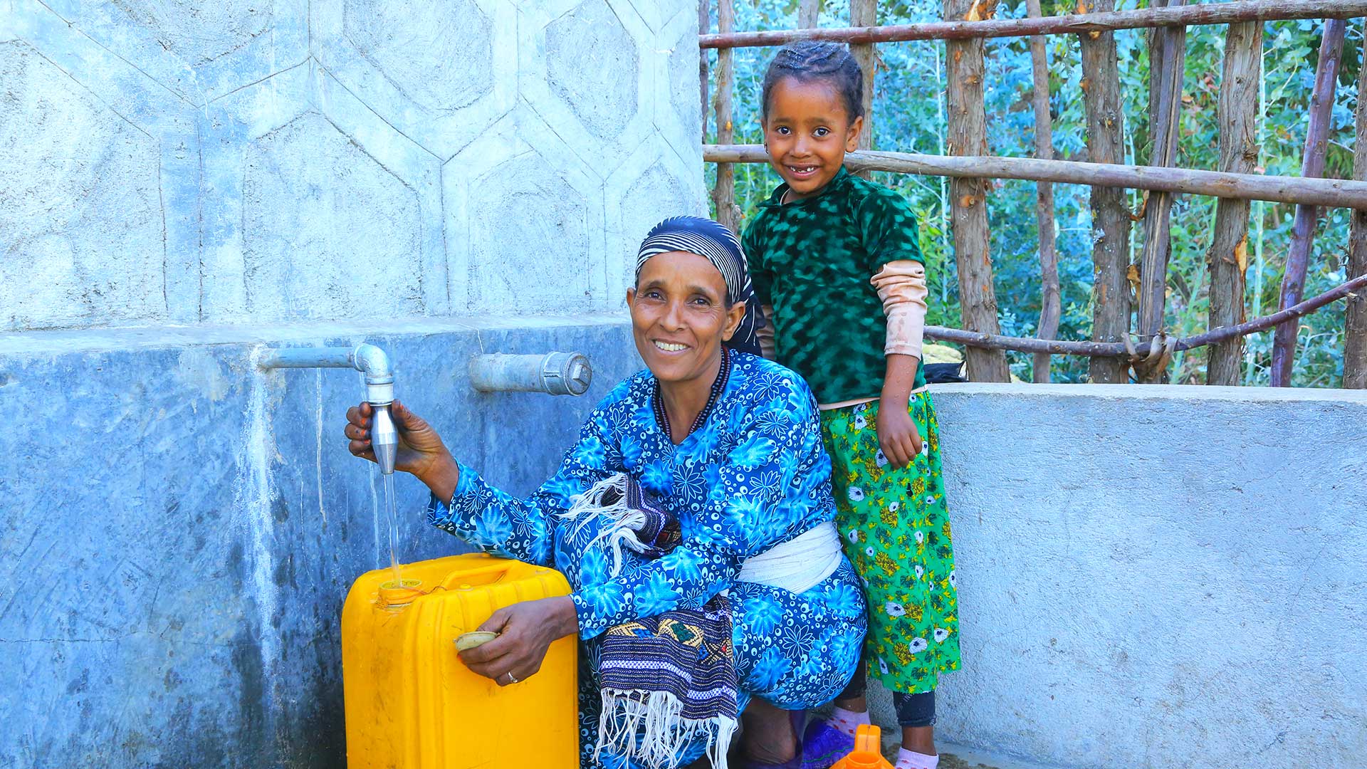 Eine Frau und ihr Kind sitzen an einer Quellfassung und lassen klares Trinkwasser in gelbe Kanister fließen.