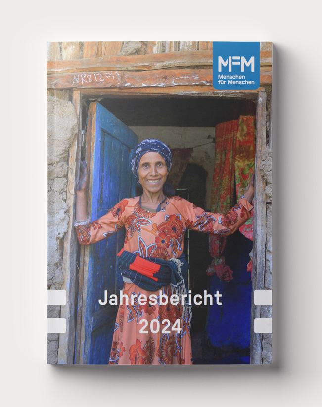 Cover des Jahresberichts 2024, das eine Frau zeigt, die in der Tür eines Hauses aus Holz und Lehm steht und lächelt.