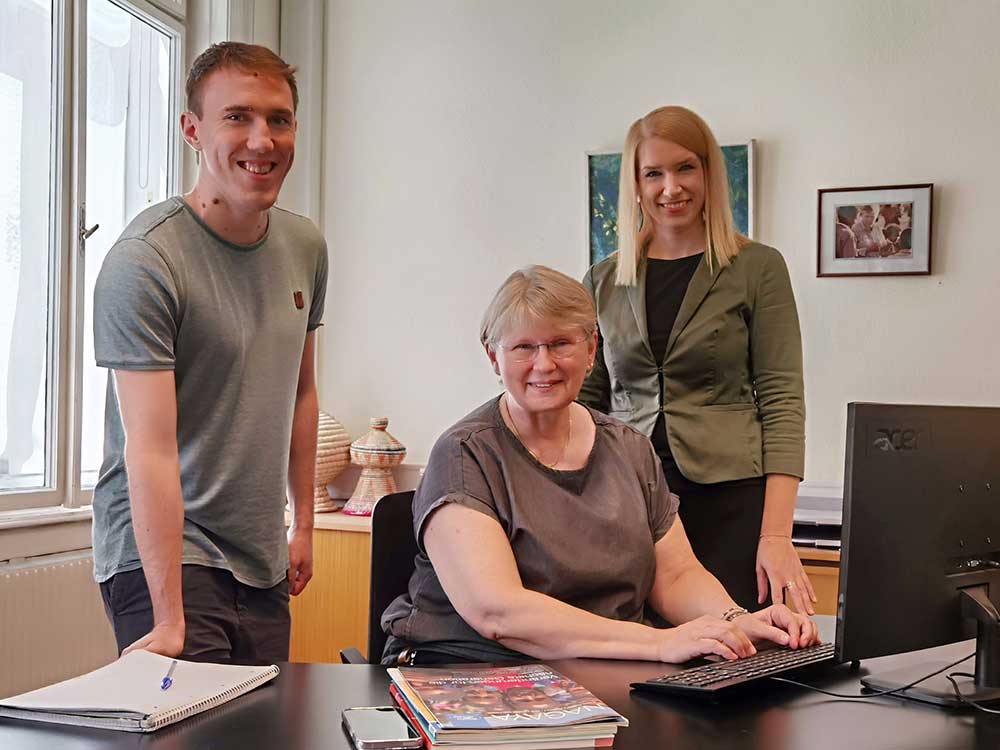Ein Bild vom Spendenservice-Team von Menschen für Menschen. Zwei Frauen und ein Mann sitzen bzw. stehen an einem Schreibtisch und lächeln in die Kamera