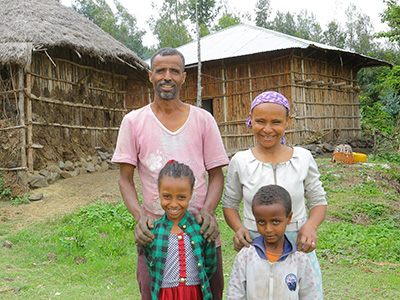 Glückliche Familie mit Vater, Mutter, Tochter und Sohn, steht vor traditionellen Hütten in einem ländlichen Dorf in Äthiopien, umgeben von grüner Natur.