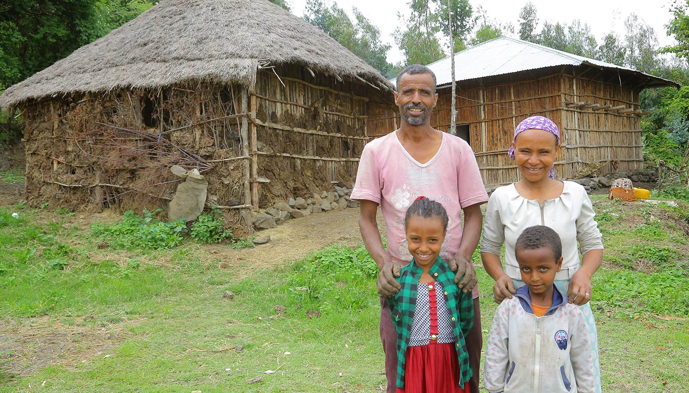 Glückliche Familie mit Vater, Mutter, Tochter und Sohn, steht vor traditionellen Hütten in einem ländlichen Dorf in Äthiopien, umgeben von grüner Natur.